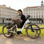 Man sitting on a bench with a bicycle, reading a newspaper in an urban setting.